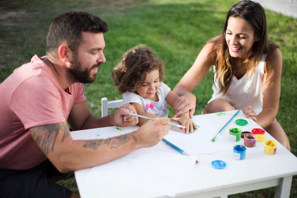 A family enjoys a painting activity on a sunny day, with colorful paint pots and brushes on a white table in a grassy outdoor setting.