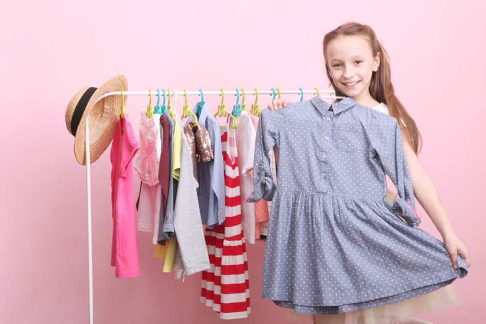 A girl holds up a blue polka dot dress in front of a colorful clothing rack against a pink backdrop. A straw hat hangs nearby.