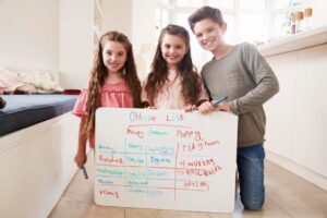 Three children stand together holding a chore list on a large whiteboard, with tasks outlined for each day of the week in colorful markers.