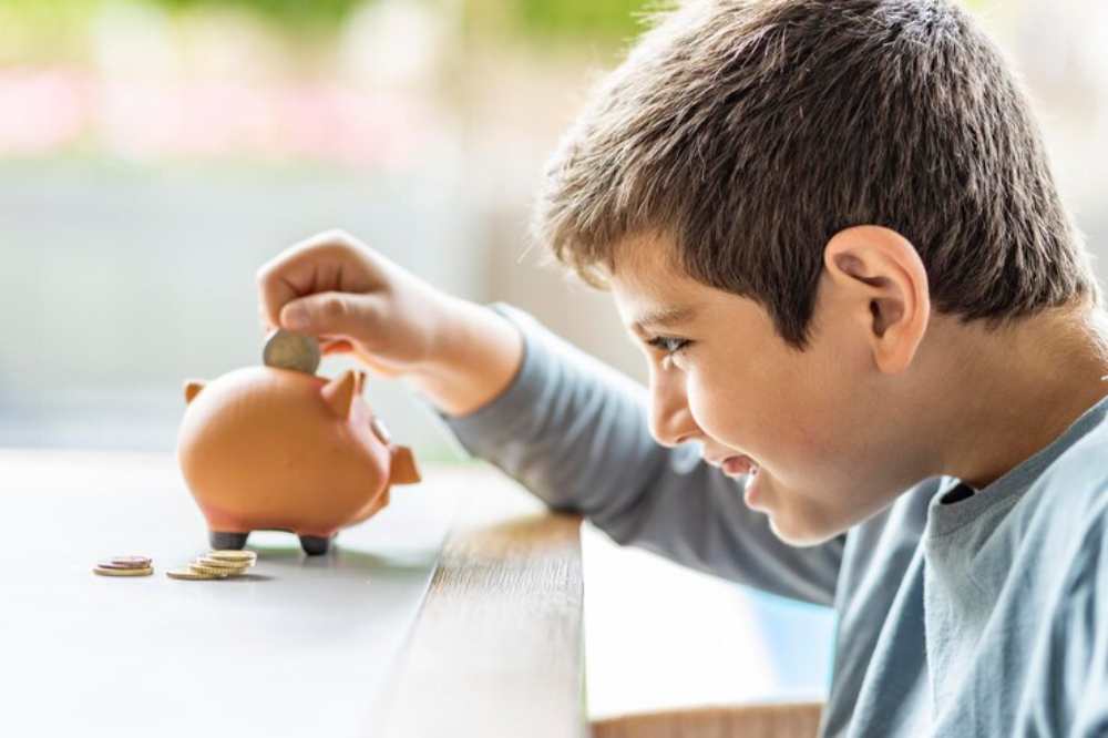 A child places a coin into a piggy bank on a table, with additional coins scattered nearby and soft light filtering in from outside.
