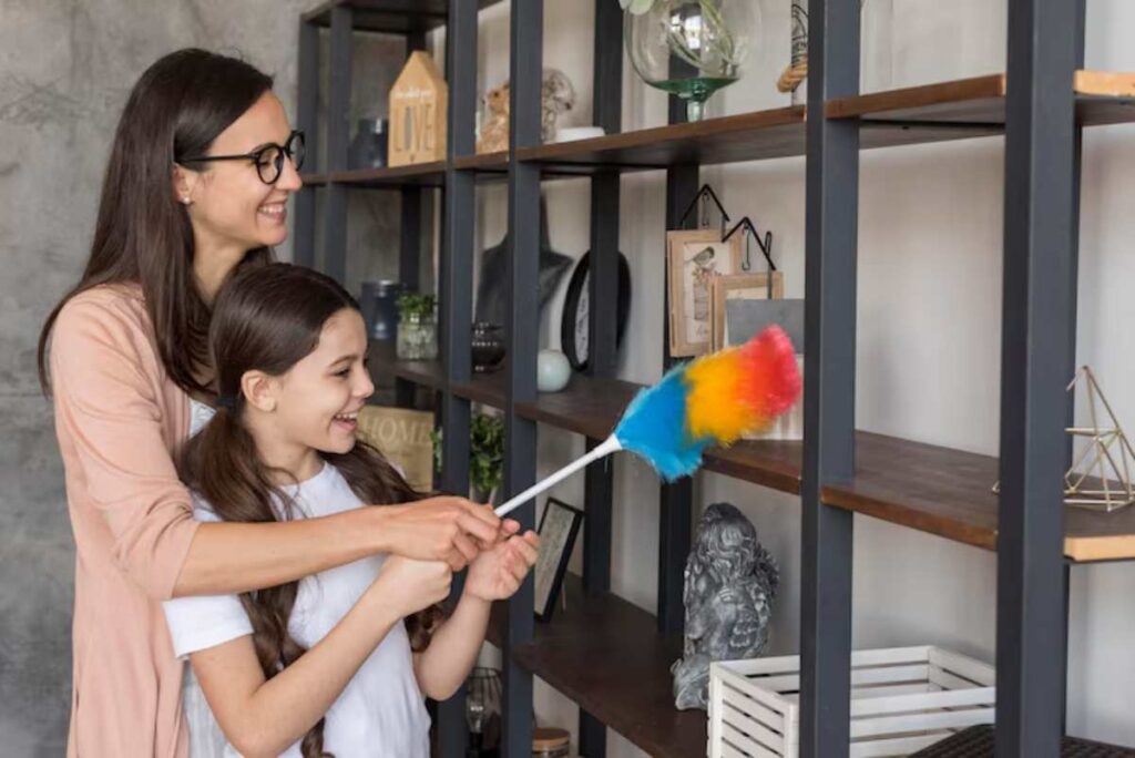 A woman and a girl clean shelves adorned with decorative items, using a colorful duster in a modern, minimalistic interior.