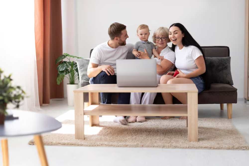 A family of four gathers on a couch, engaging with a laptop on a coffee table, surrounded by indoor plants and warm lighting.