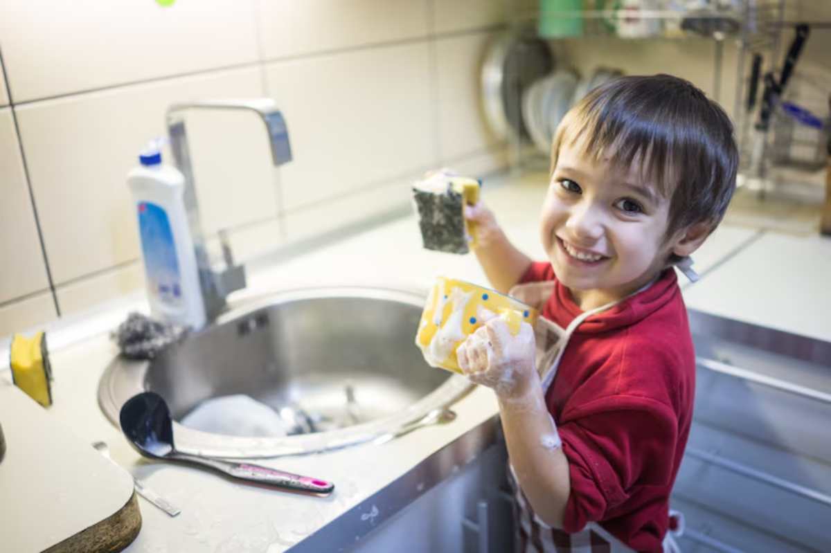A child in a red shirt and apron holds two sponges, surrounded by soapy water at a kitchen sink, engaged in washing dishes.