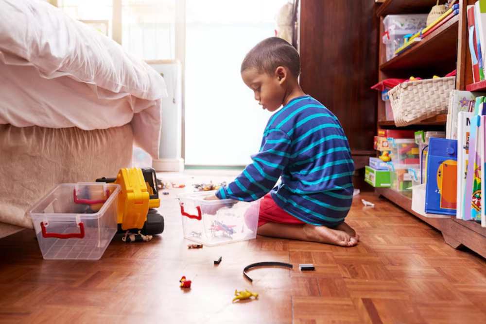 A child in striped pajamas sits on the floor, sorting toys into clear bins amidst a colorful, toy-filled room.