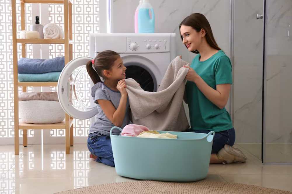 A child and an adult sort laundry together by a washing machine, with a basket filled with colorful clothes beside them.