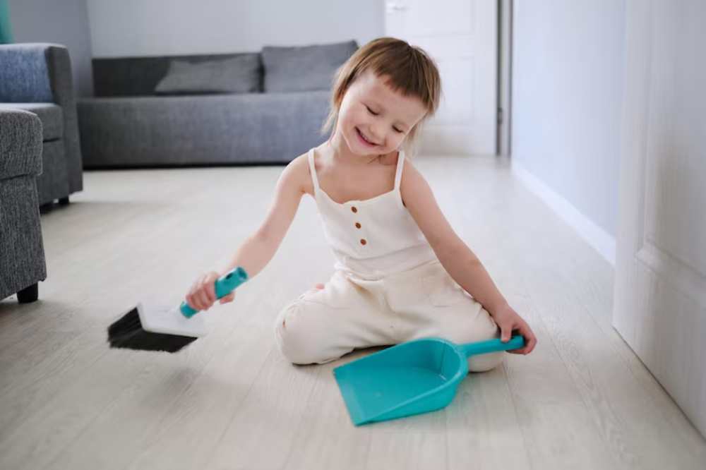 A young child sits on the floor, using a dustpan and brush to clean the light wooden surface in a cozy living space.