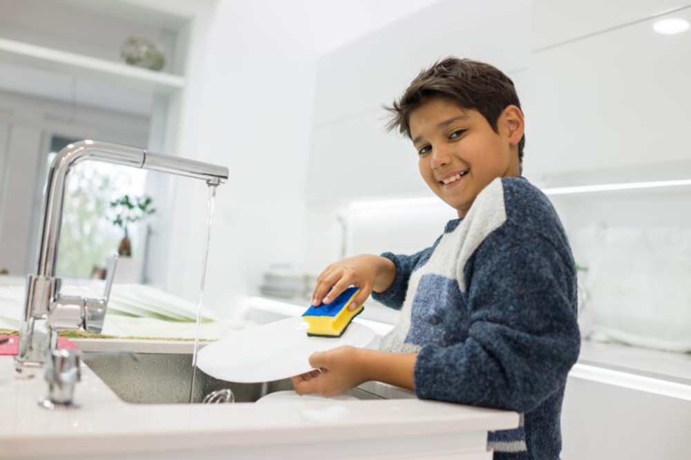 A child wearing a cozy sweater washes a white plate at a modern kitchen sink, with water running and a bright, airy atmosphere.