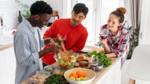 Three siblings enjoy a cooking session in a bright kitchen, surrounded by fresh vegetables and fruits, sharing laughter and drinks.