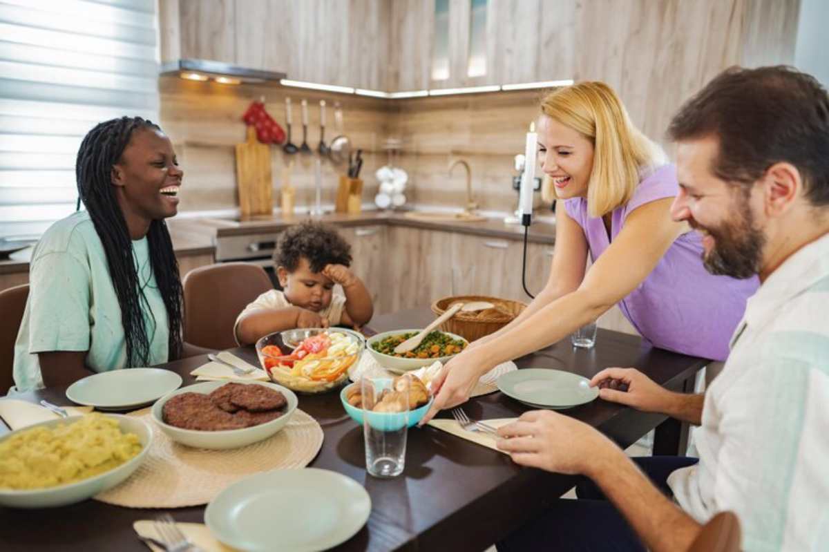 A family gathers around a dining table filled with various dishes, including salads, bread, and burgers, enjoying a meal together.
