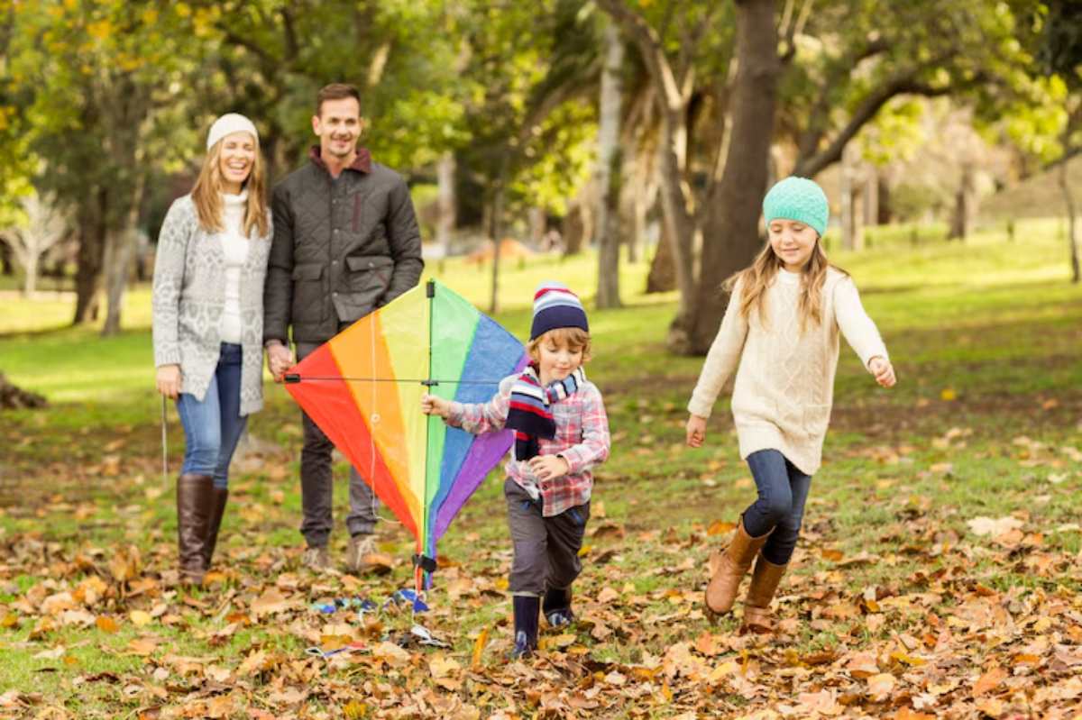 A family enjoys a sunny day in a park, flying a colorful kite among fallen leaves and trees, with children playing happily.
