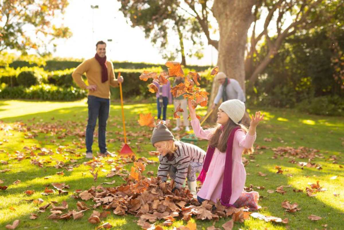 Children play joyfully in a sunlit park, surrounded by piles of autumn leaves, while adults tidy up in the background.