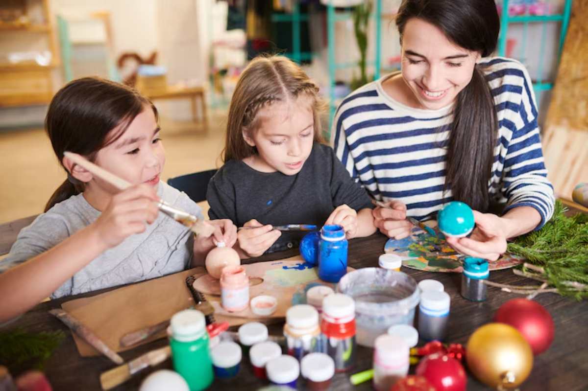 Children and an adult engage in a creative activity, painting eggs and ornaments with various colors and materials on a table.