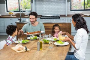 A family enjoys a meal together at a wooden dining table, featuring dishes of salad, meat, and pasta in a bright kitchen setting.