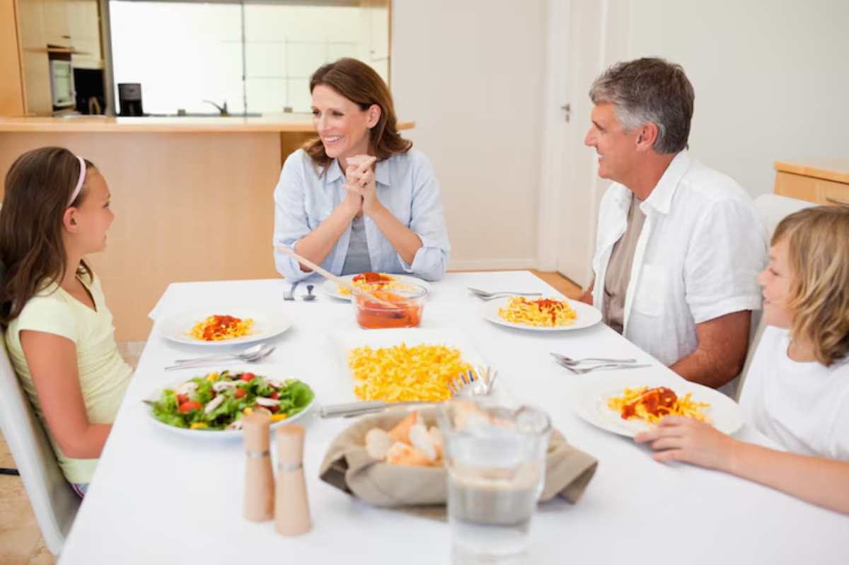A family gathers around a dining table, enjoying plates of pasta and salad, with drinks and bread in a bright, modern kitchen.