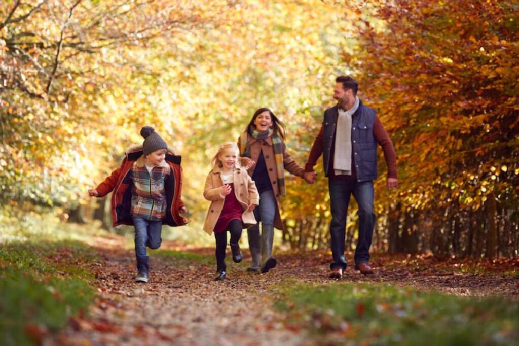 A family of four enjoys a playful walk along a leaf-covered path, surrounded by vibrant autumn foliage.