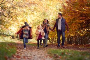 A family of four enjoys a playful walk along a leaf-covered path, surrounded by vibrant autumn foliage.