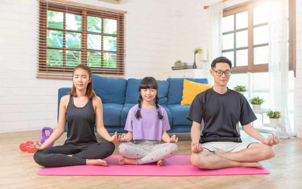 A woman, girl, and man practice meditation on a pink mat in a bright living room adorned with plants and a blue sofa.