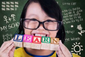 A child holds colorful wooden blocks with letters against a chalkboard featuring math equations, letters, and drawings.