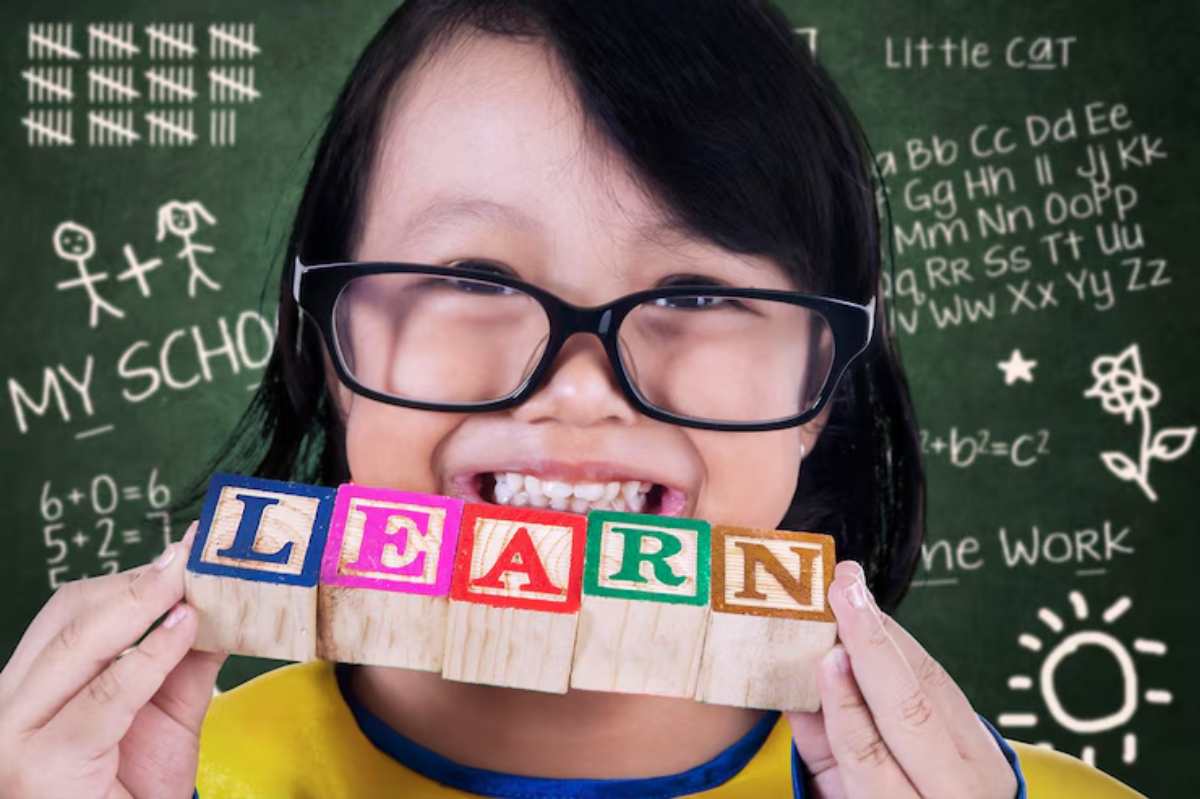 A child holds colorful wooden blocks with letters against a chalkboard featuring math equations, letters, and drawings.