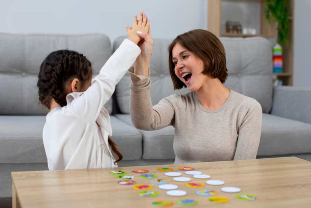 A child and an adult play a game, raising their hands in victory, with colorful game pieces scattered on the table between them.