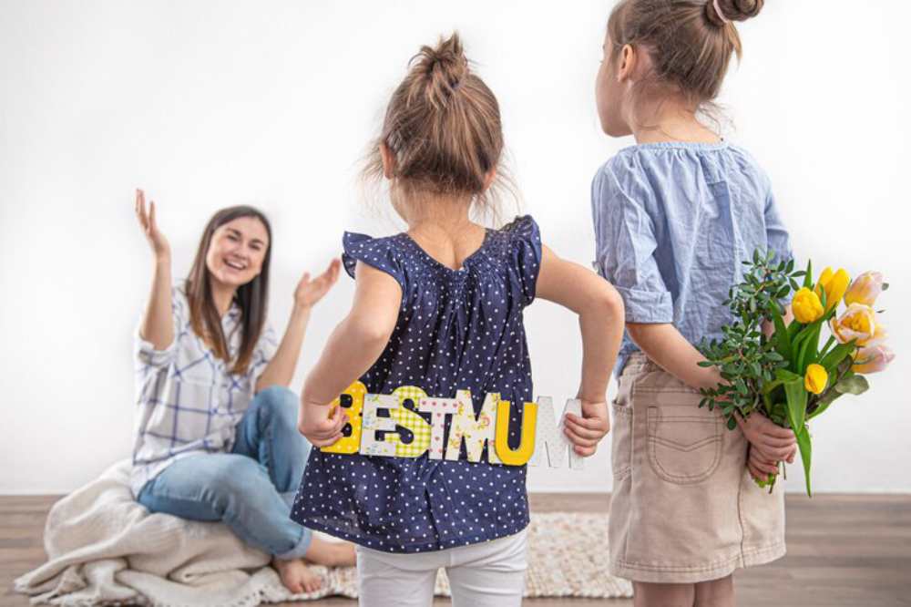 Two young girls are preparing a surprise for their mother, one holding a bouquet of flowers and the other displaying a sign that reads BEST MUM.
