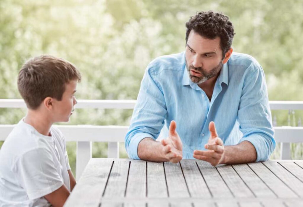 A man in a light blue shirt engages in conversation with a boy at a wooden table, surrounded by soft natural light.