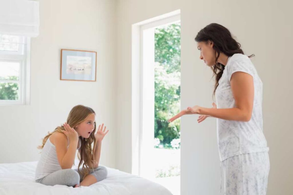A woman gestures emphatically while speaking to a girl sitting on a bed, both in a bright, airy bedroom.