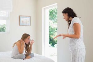 A woman gestures emphatically while speaking to a girl sitting on a bed, both in a bright, airy bedroom.