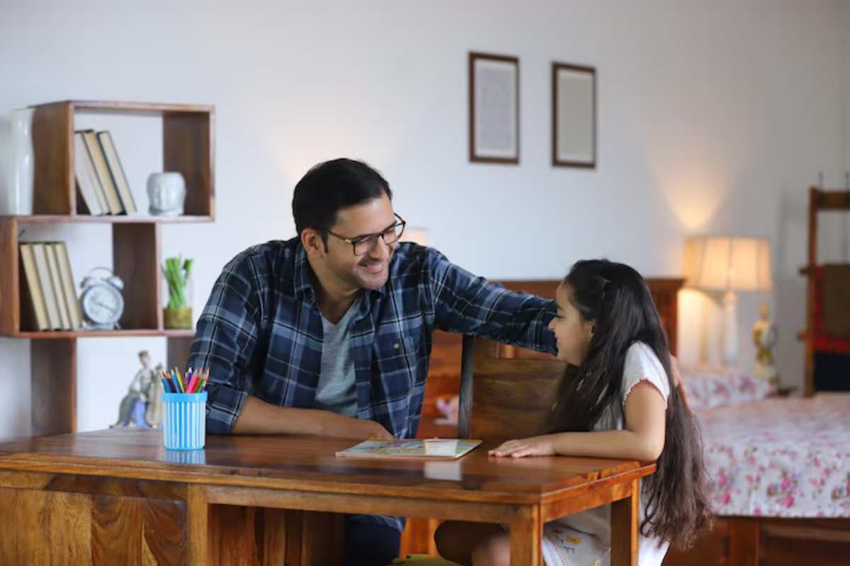 A parent and child sit together at a wooden table, engaging in conversation with art supplies and books nearby. Soft lighting illuminates the room.