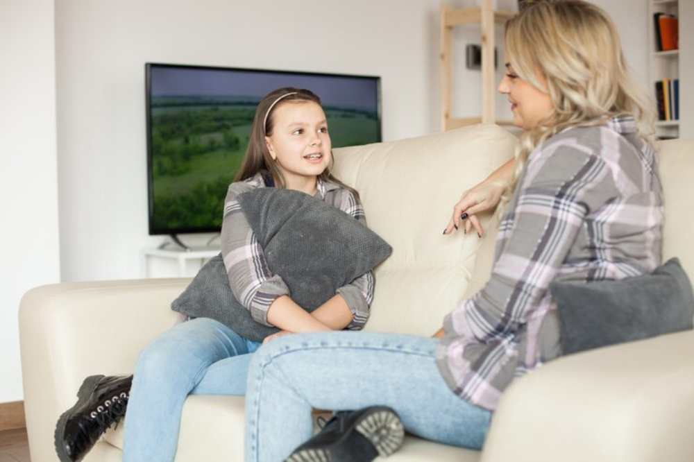A girl and woman in plaid shirts sit on a beige couch, engaged in conversation, with a scenic television landscape in the background.