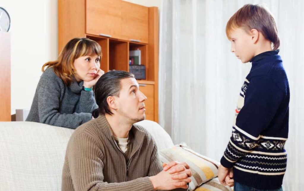 A young boy stands, looking at a man seated on a couch, while a woman observes from behind in a cozy living room setting.