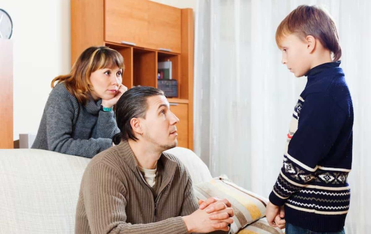 A young boy stands, looking at a man seated on a couch, while a woman observes from behind in a cozy living room setting.