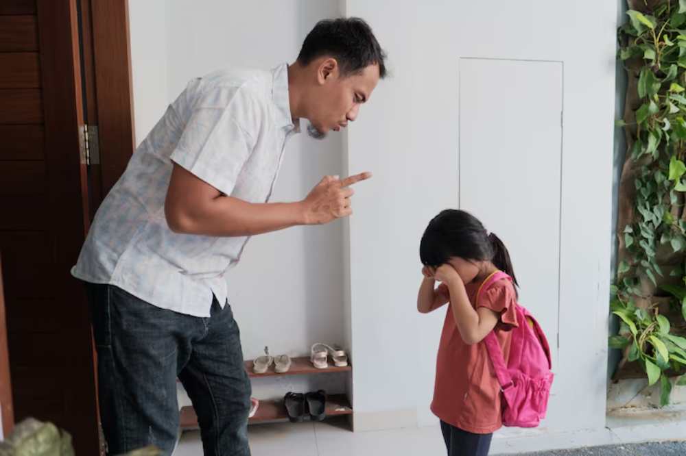 A parent scolds a young girl in a pink shirt and backpack, who is covering her face with her hands, standing in a modest home entryway.