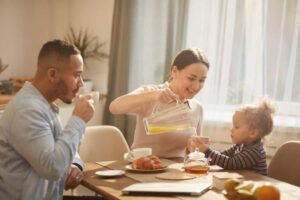 A woman pours orange juice for a child at a cozy breakfast table, surrounded by food and drinks in a sunlit room.