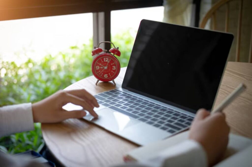 A person works on a laptop at a wooden table, with a red alarm clock nearby and green foliage visible through the window.