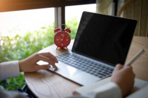 A person works on a laptop at a wooden table, with a red alarm clock nearby and green foliage visible through the window.
