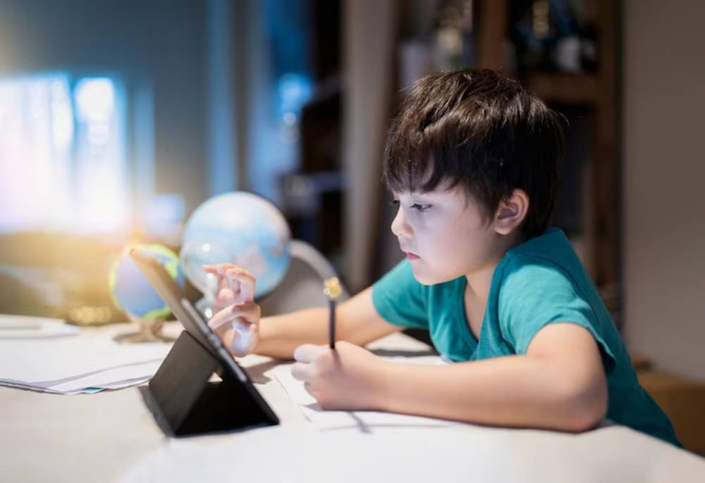 A child in a turquoise shirt is studying at a desk with a tablet, surrounded by books and a globe, focusing intently on their work.