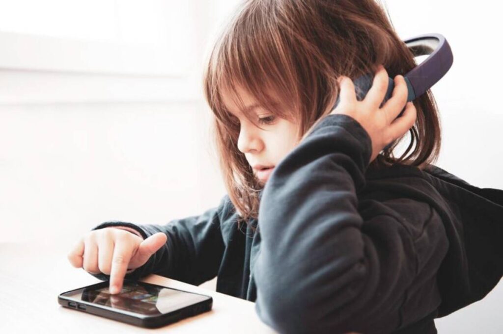 A child wearing headphones interacts with a tablet, focused on the screen, seated at a table near a bright window.