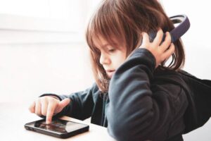 A child wearing headphones interacts with a tablet, focused on the screen, seated at a table near a bright window.