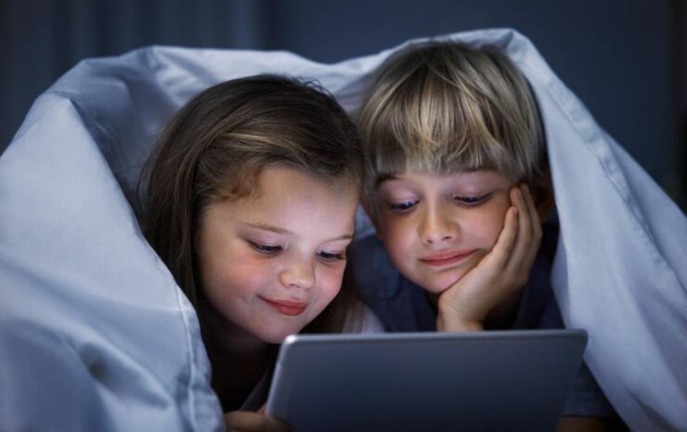 Two children under a white blanket, closely watching a tablet screen in a dimly lit room.