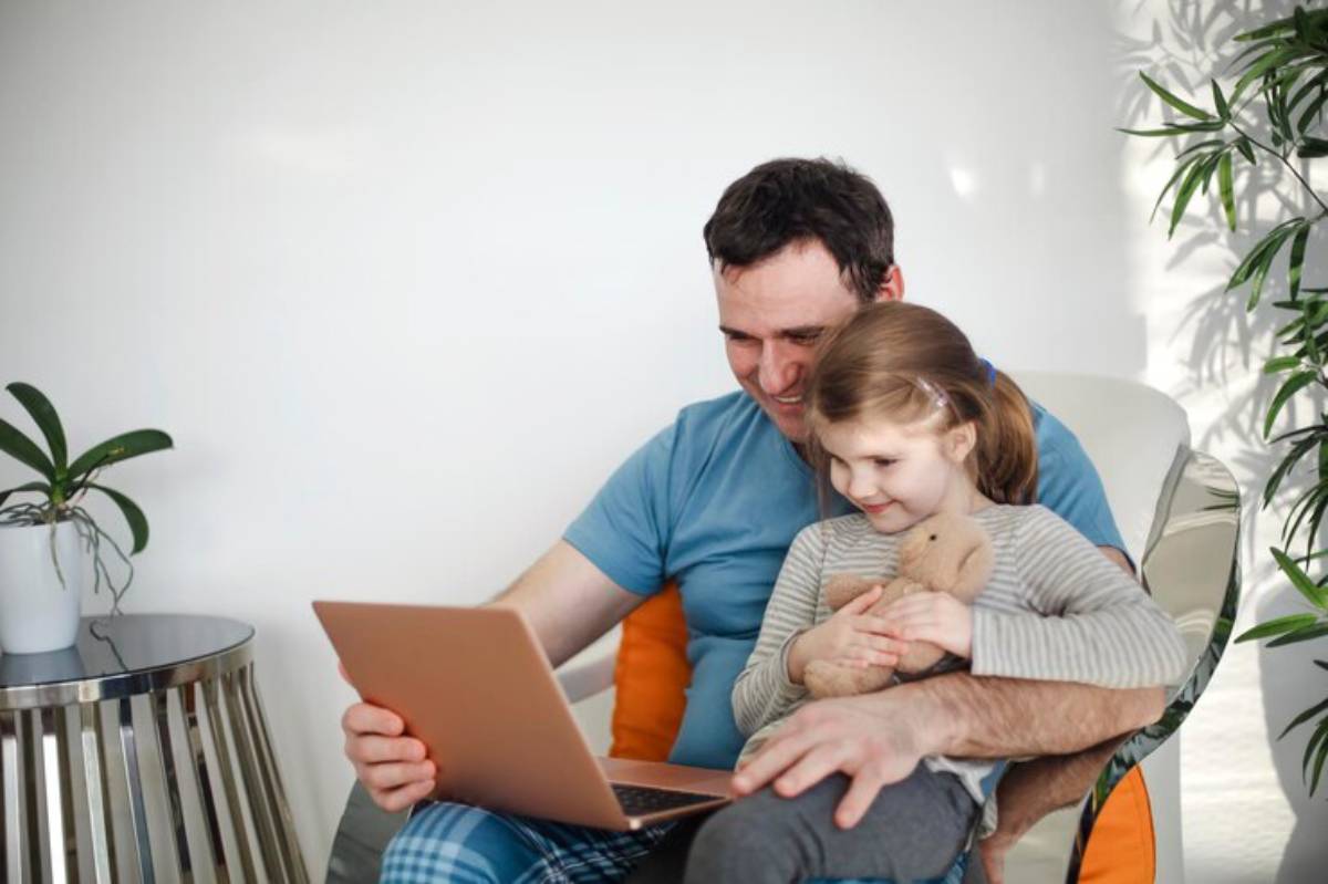 A parent and child sit together on a chair, focused on a laptop, with a cozy indoor setting and a potted plant nearby.