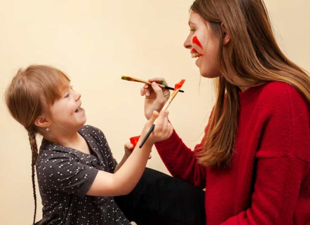 A young girl in a polka-dot shirt paints a woman's face with colorful brushes, showcasing a playful art session against a cream background.