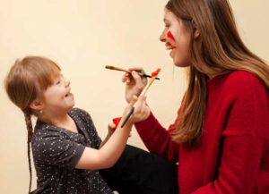 A young girl in a polka-dot shirt paints a woman's face with colorful brushes, showcasing a playful art session against a cream background.