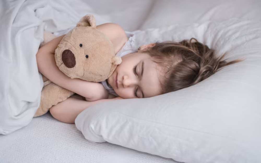 A young girl peacefully sleeps on a white bed, hugging a teddy bear tightly against her chest. Soft pillows surround her.