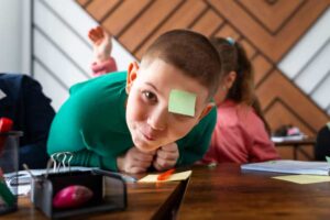 A child in a green sweater leans over a wooden table with colorful stationery while classmates engage in activities in the background.