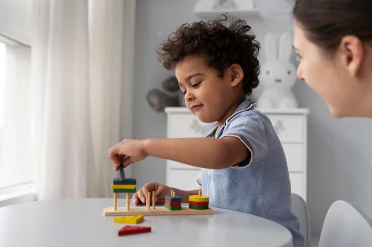 A child plays with colorful stacking toys on a table, focusing intently on arranging wooden pieces. Sunlight streams through nearby curtains.