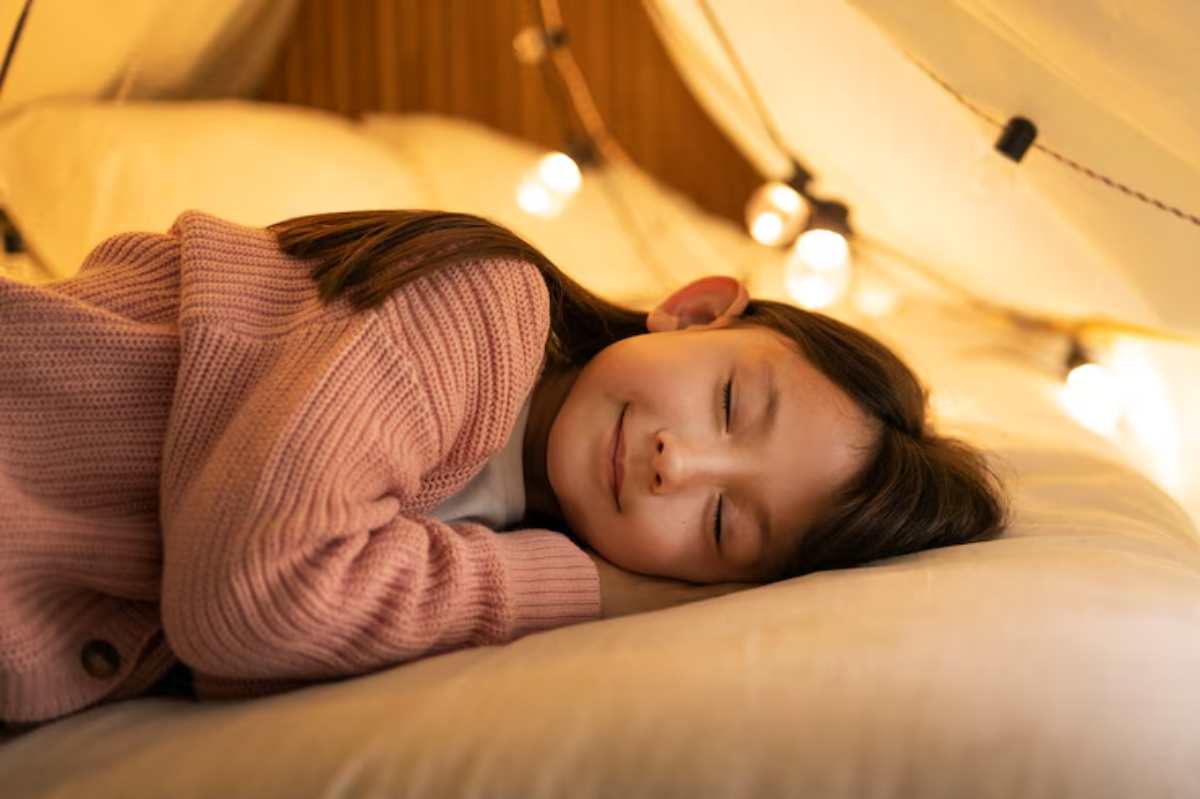 A child lies comfortably on a bed with soft white blankets, wearing a cozy pink cardigan, surrounded by warm, glowing fairy lights.