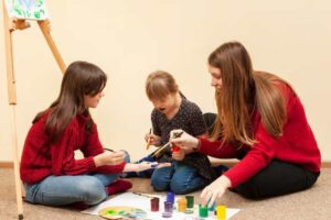 Three children are engaged in a creative painting activity on the floor, surrounded by colorful paint pots and art supplies.