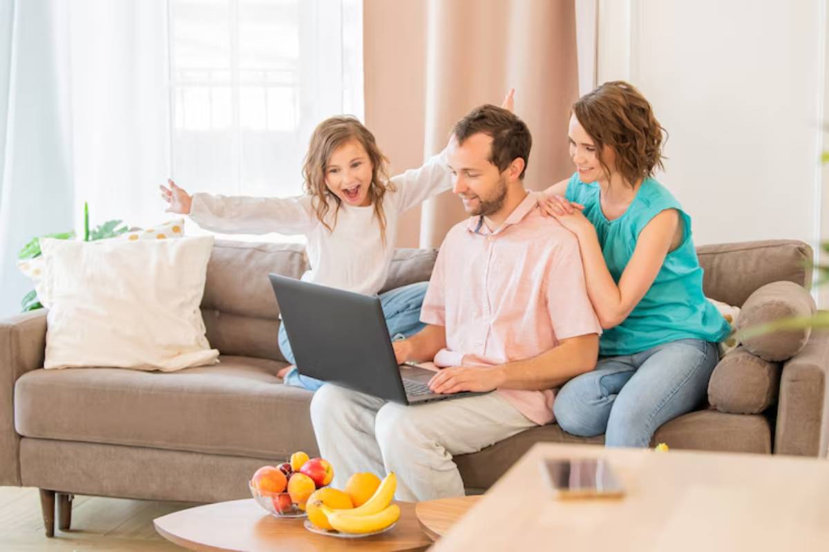 A family sits on a couch with a laptop, a child excitedly raising hands behind them, while a bowl of fruit rests on a nearby table.