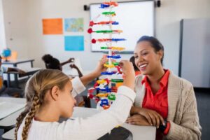 A child assembling a colorful model with plastic pieces in a classroom, while an adult observes attentively in the background.